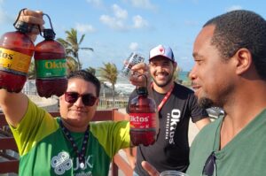 Travel Jones standing with the manager of Oktos Brewery and a tour guide on the patio overlooking the ocean, showcasing a selection of the brewery's finest beers.